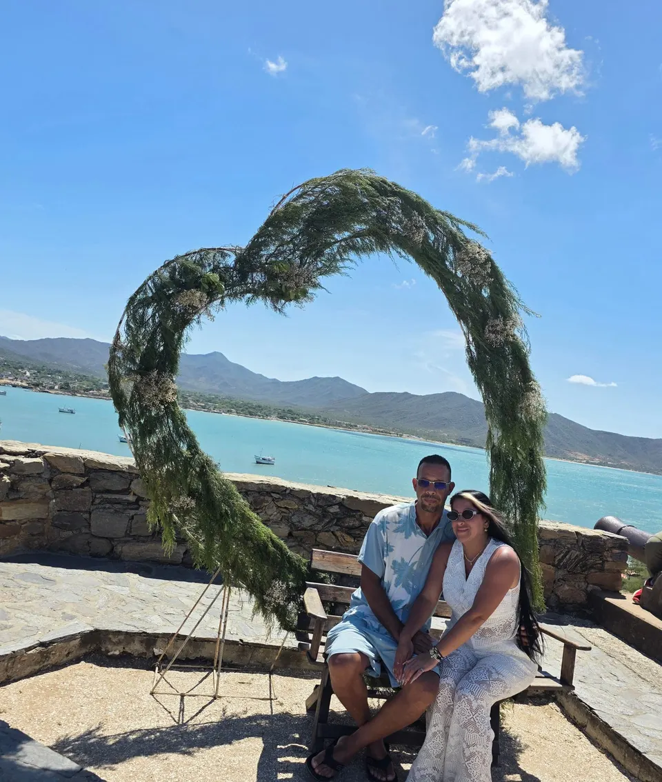 Pareja feliz posando bajo arco de corazón con vistas al mar turquesa y montañas en Isla Margarita.